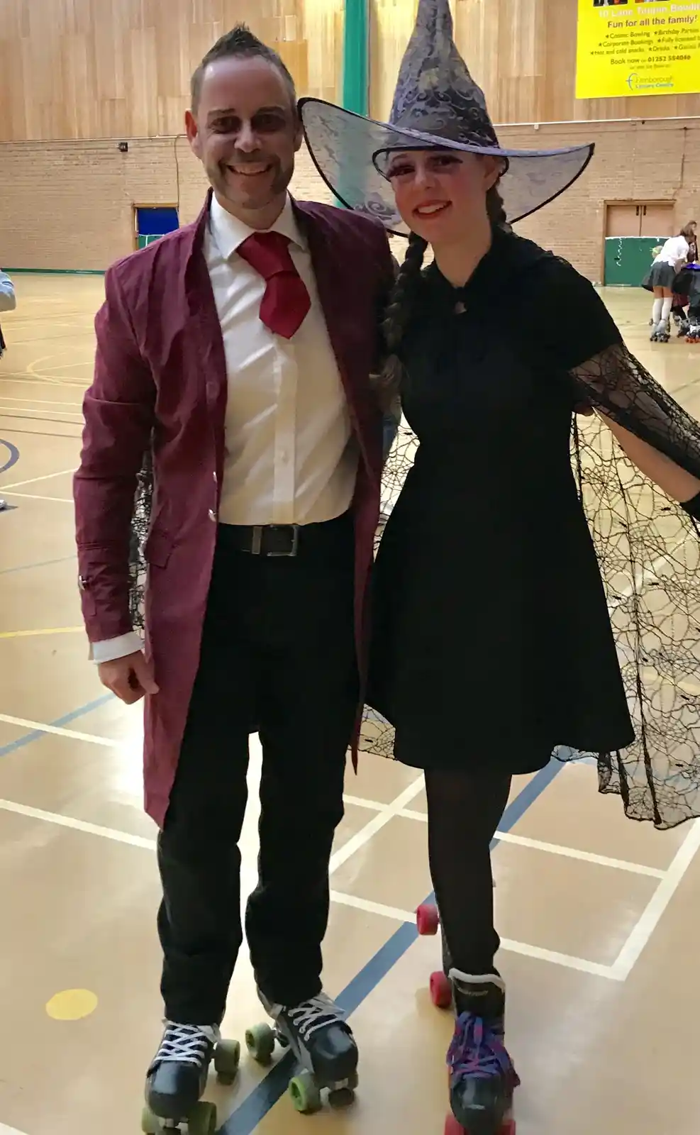 Two people in costumes pose together on roller skates inside a gymnasium. The man wears a maroon coat, white shirt, red tie…