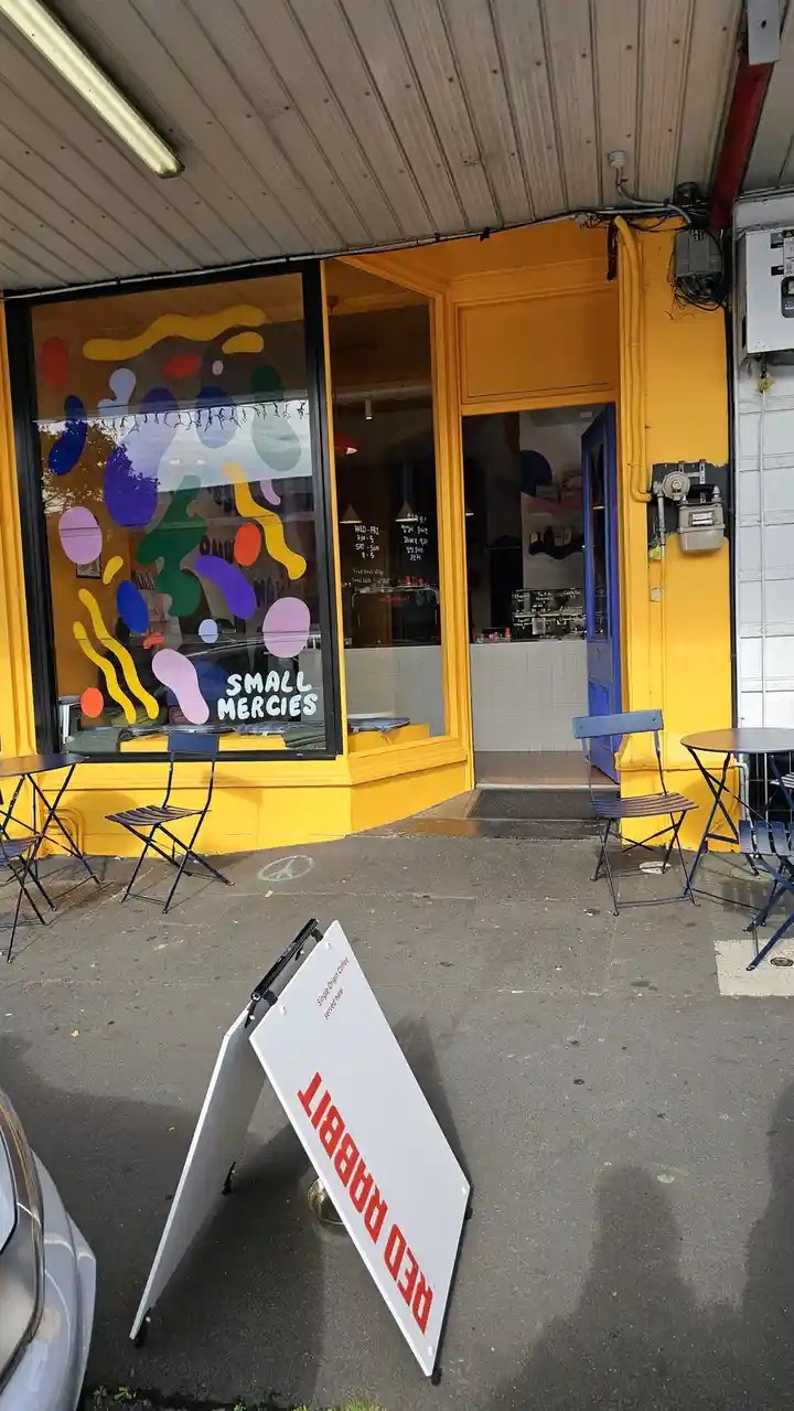 A photo of a bright yellow cafe storefront with colorful abstract decals on the window and outdoor seating. A white A-frame…