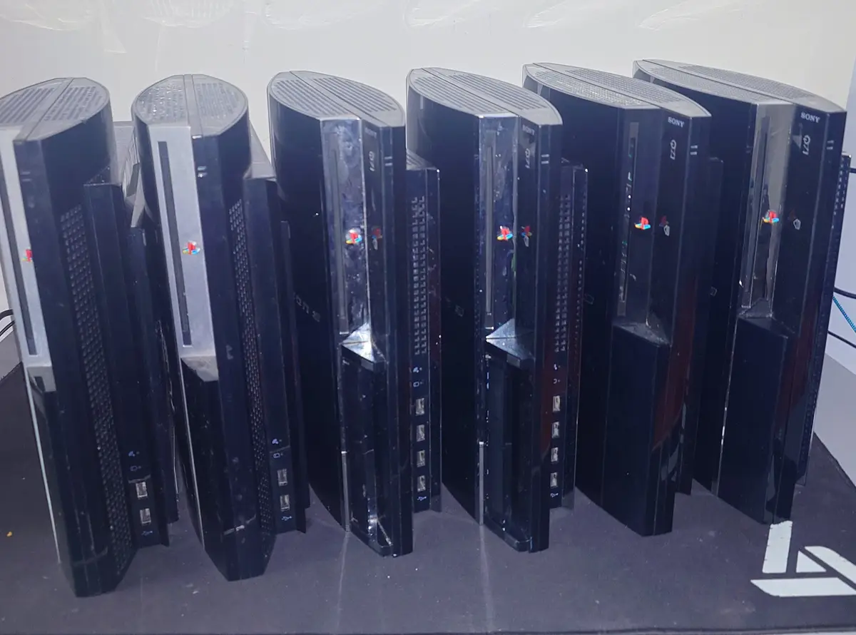 A row of six PlayStation 3 consoles stands upright on a dark surface, angled slightly inward. Each black console features…