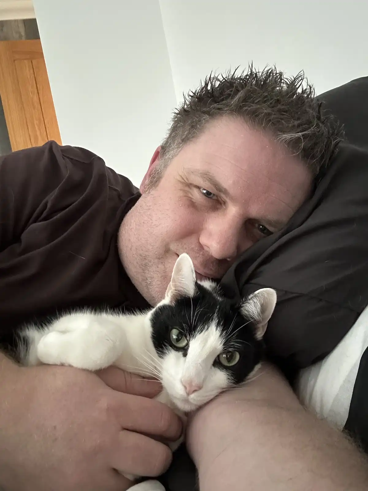 A man with messy hair and a brown shirt lies in bed, smiling slightly while holding a black-and-white cat that looks…