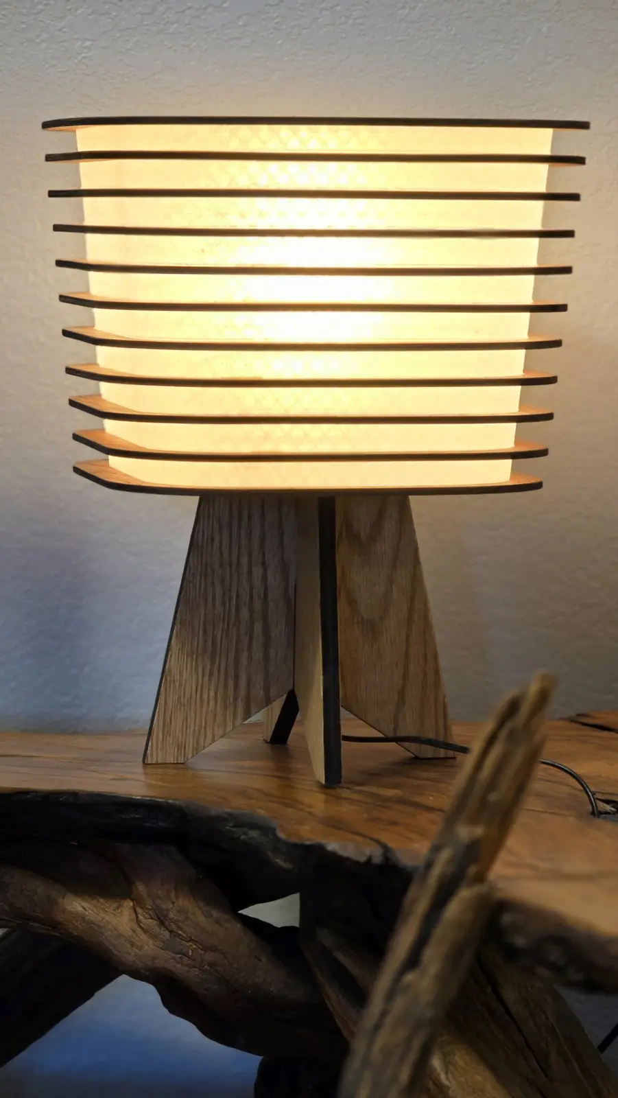 A close-up photo of a modern wooden table lamp with horizontal slats glowing warmly, sitting on a rustic wooden table. The…