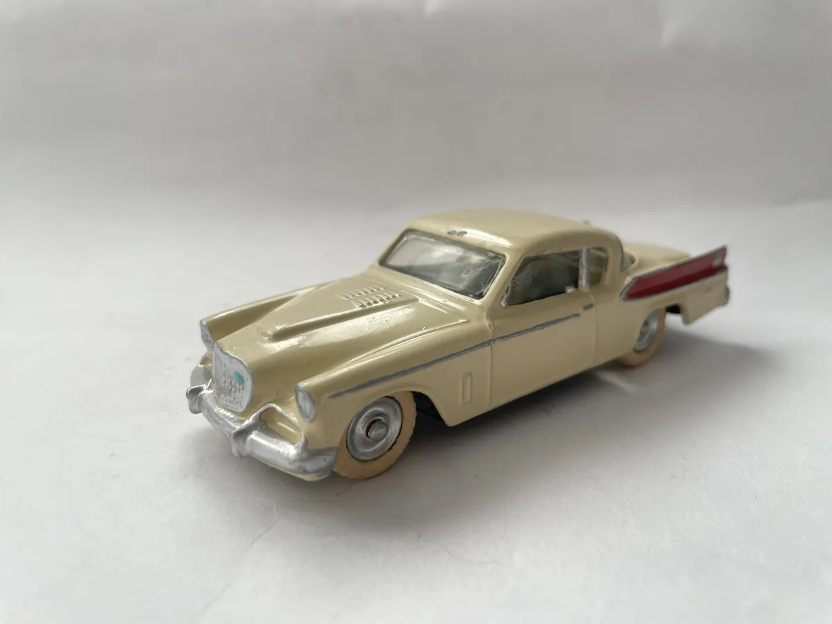 A beige vintage-style toy car with silver trim and red tail fin detail sits on a plain white surface. The car appears…