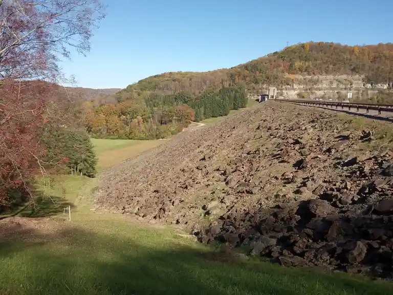 A wide shot of a large, rocky dam or embankment in autumn, with colorful trees on surrounding hills under a clear blue sky…