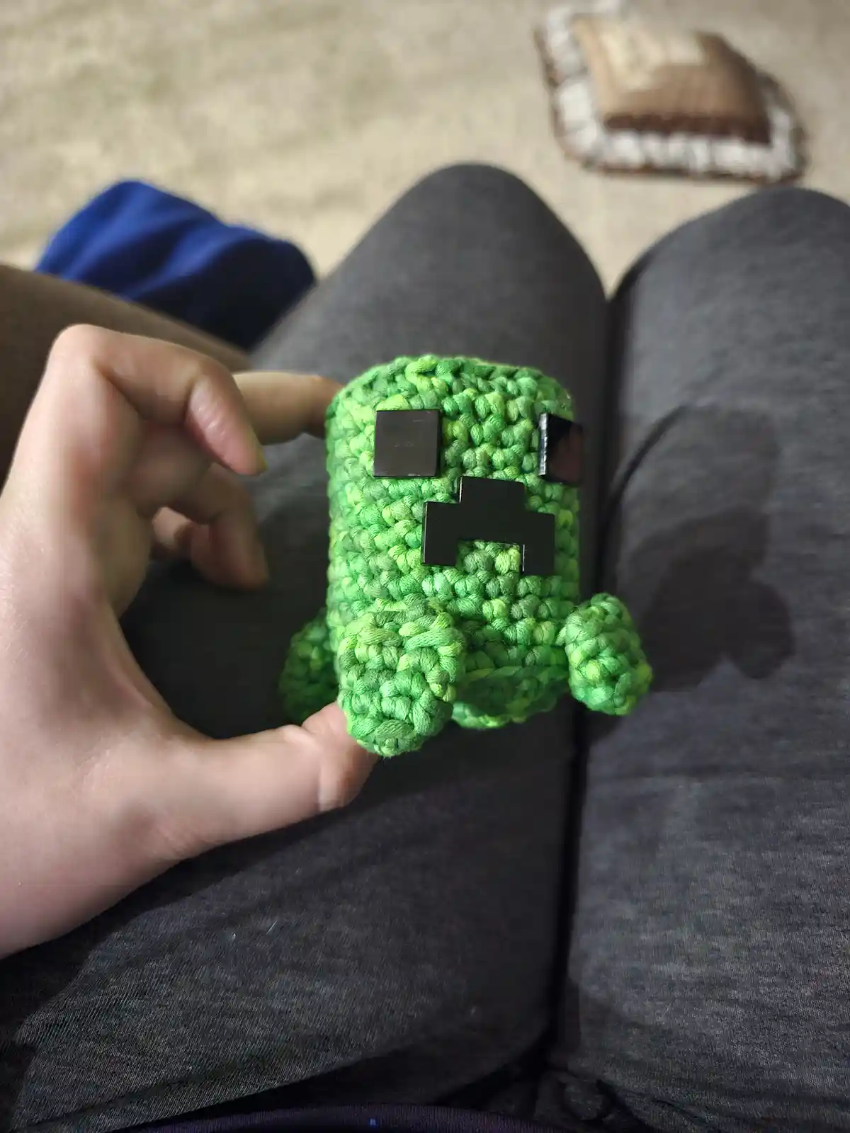 A hand holds a small, green, crocheted plushie shaped like a Minecraft creeper, featuring black square eyes and mouth. The…