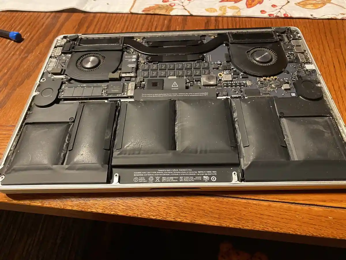 A disassembled MacBook Pro lies open on a wooden table, exposing its internal components including two cooling fans, circuit…