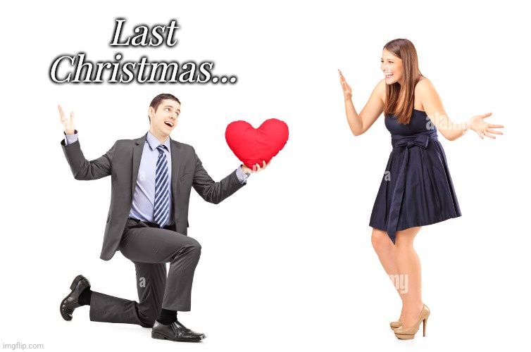 A man in a suit kneels holding a red heart-shaped pillow while looking up with a hopeful expression. A woman in a dark dress…