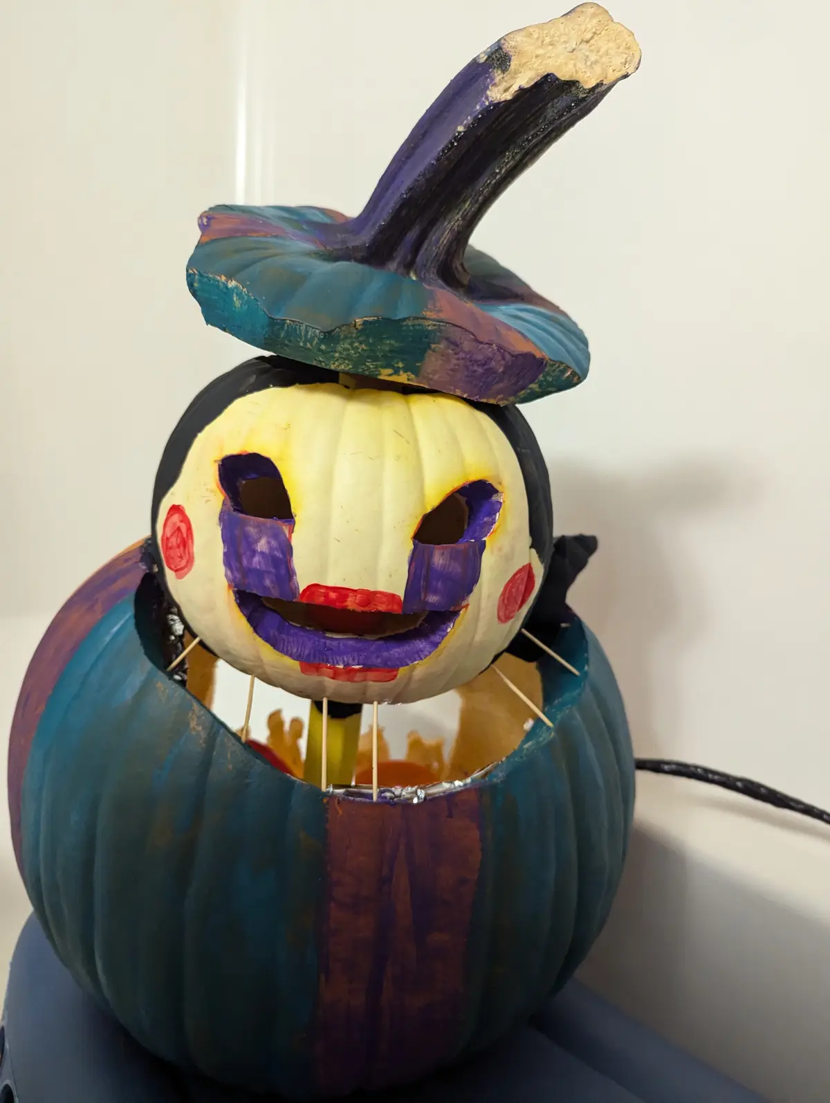 A creatively carved and painted Halloween pumpkin sculpture, featuring a smaller pumpkin head with a painted clown-like face…