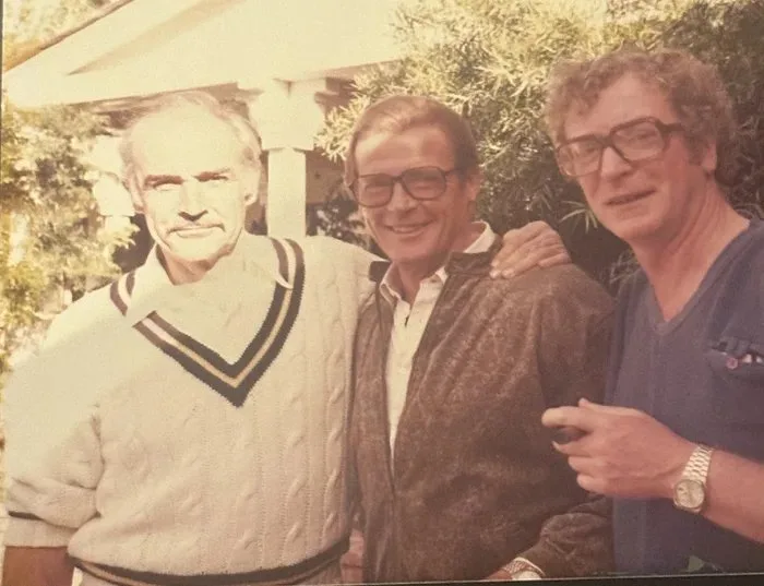 A vintage, slightly faded photograph shows three men posing outdoors. The man on the left, with white hair and a cable-knit…