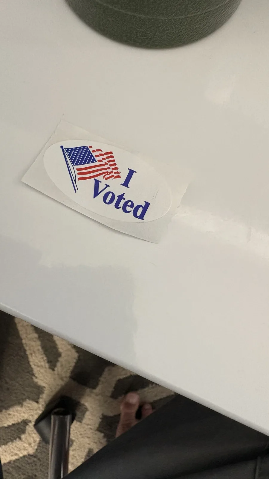 A close-up photo shows a slightly crumpled 'I Voted' sticker with an American flag design stuck to a white surface, likely a…