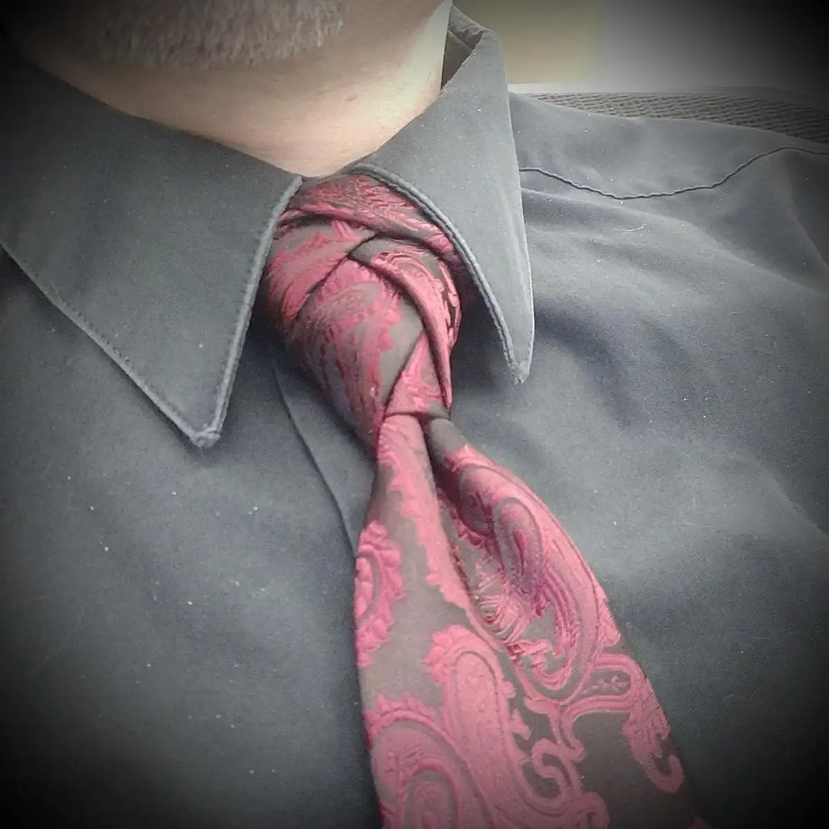 A close-up photo shows a man's neck and chest wearing a dark gray collared shirt with a red and black paisley tie that is…