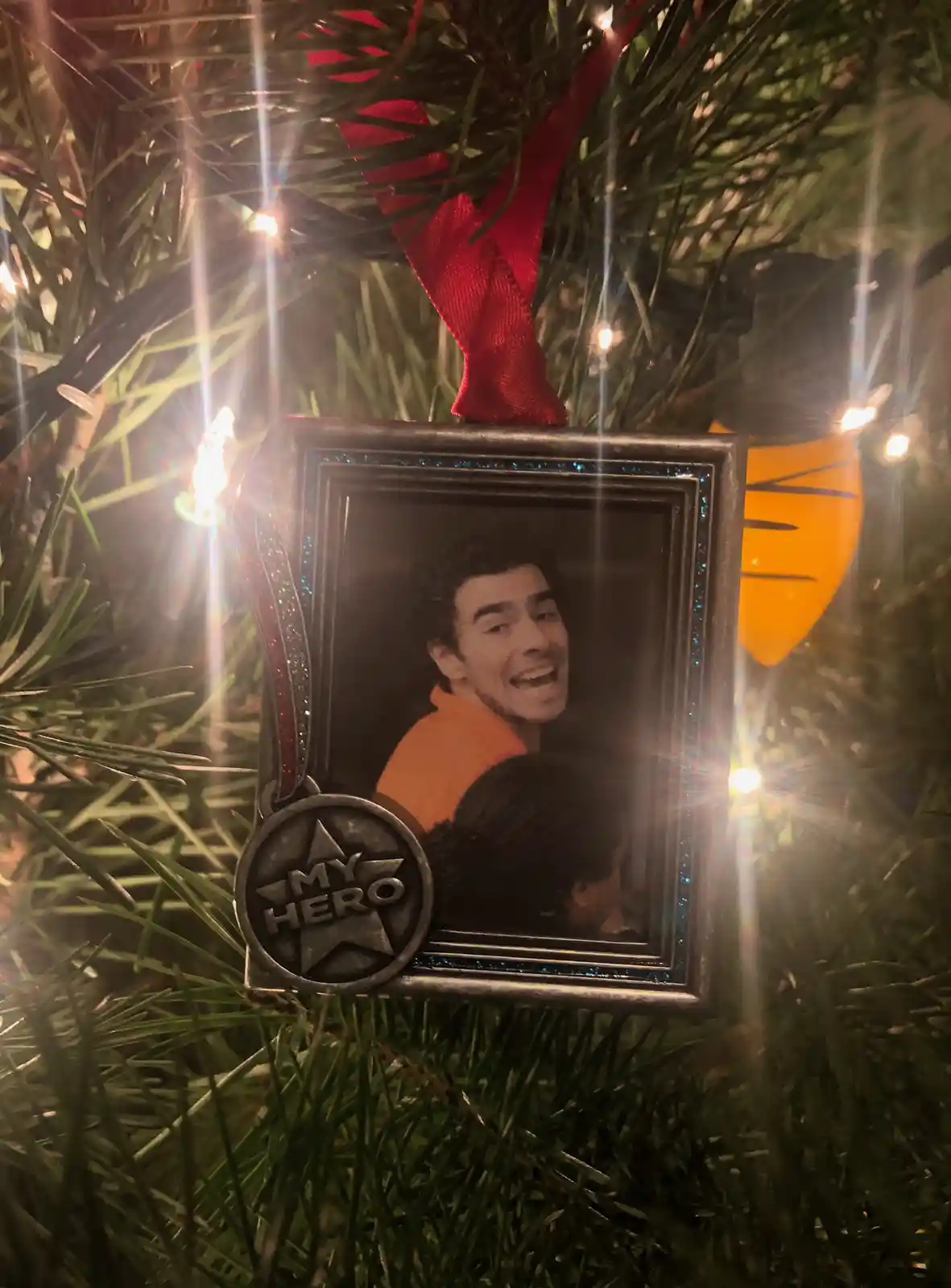 A close-up of a Christmas ornament hanging on a pine tree, featuring a framed photo of a smiling man in an orange shirt. The…