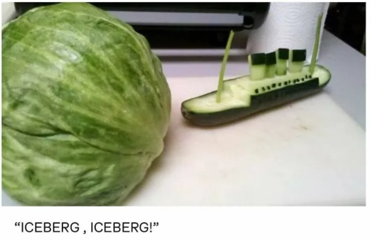 A large green cabbage sits on a white cutting board next to a cucumber carved into the shape of a ship with four…