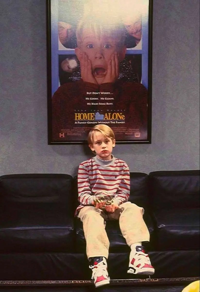A young boy in a red-and-white striped shirt and beige pants sits on a black couch, looking bored and unimpressed. Behind…