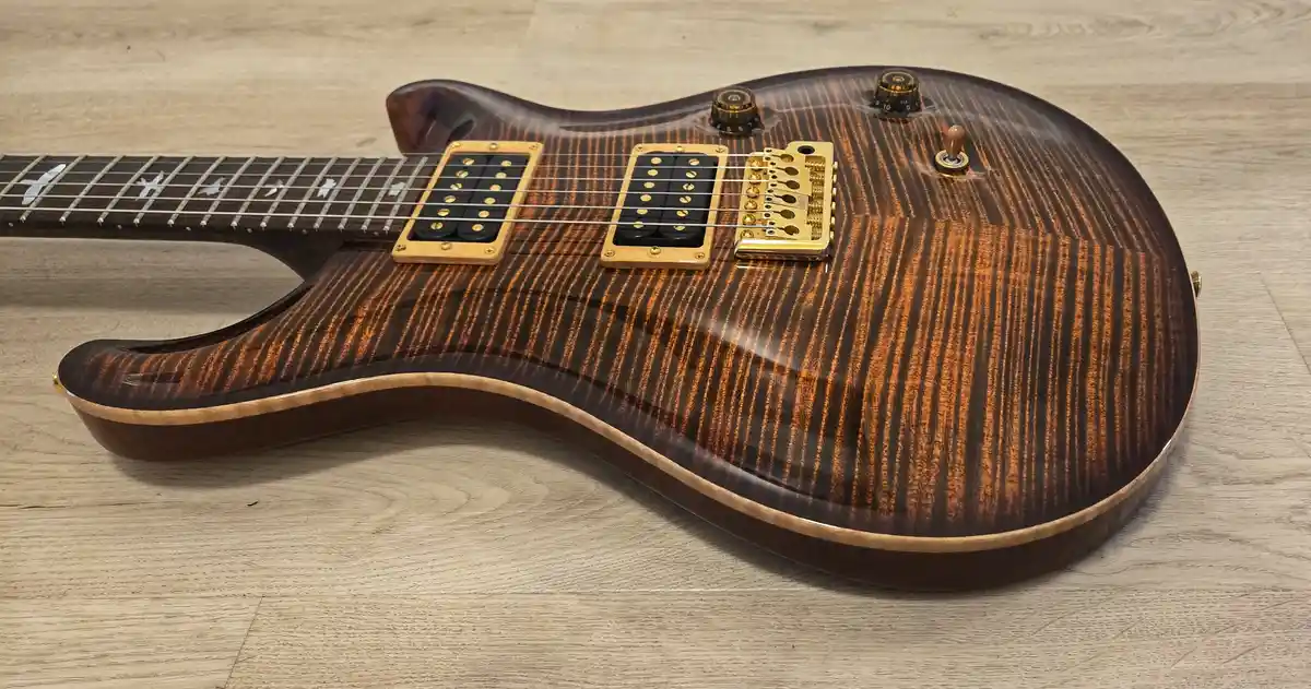 A close-up photo of a high-end electric guitar with a striking flame maple top, gold hardware, and black pickups, resting on…