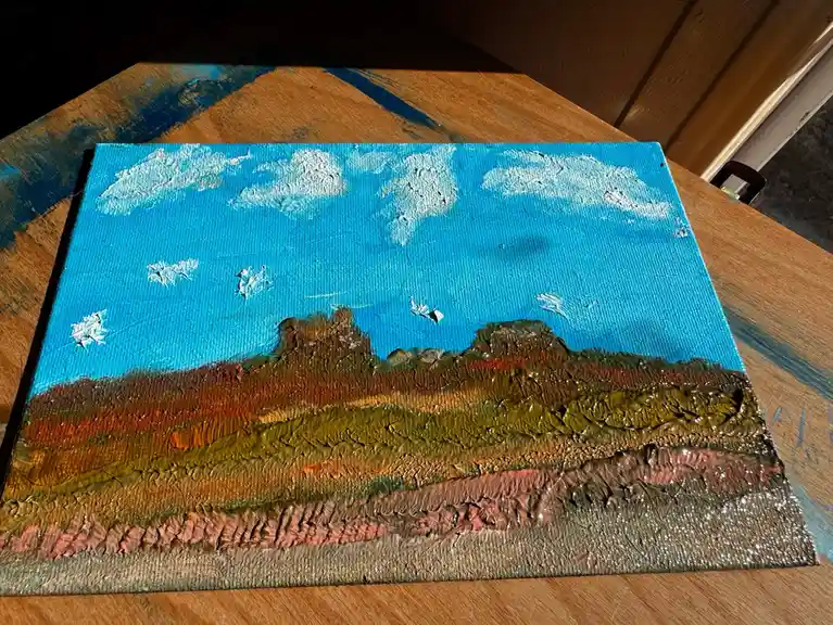 A thickly textured, impasto-style painting of a landscape on canvas, resting on a wooden table. The sky is bright blue with…