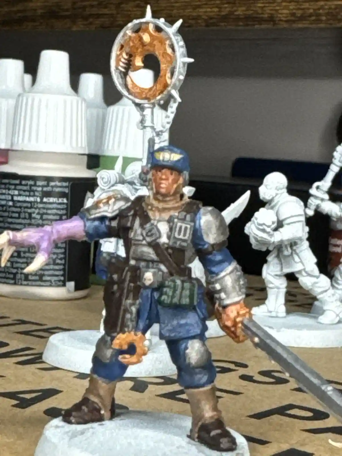 A painted miniature of a soldier in blue and silver armor stands on a base, holding a sword and a large circular weapon with…