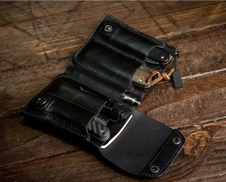a black leather knife roll with multiple slots holding various folding knives and tools, resting on a worn wooden surface…