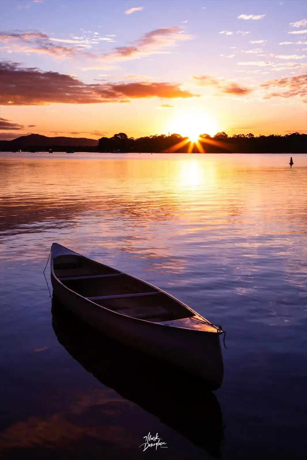 A serene sunset photograph shows a single canoe floating on calm water, with the sun setting behind distant trees and hills…
