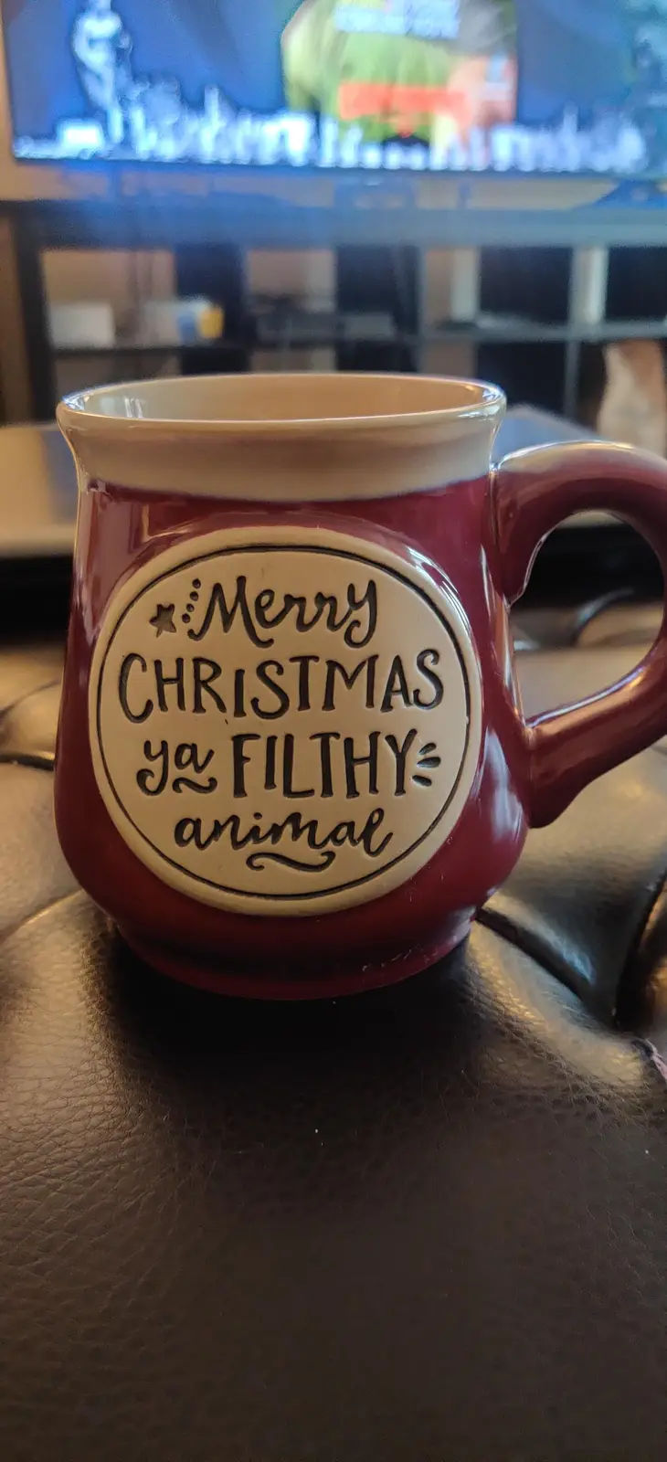 A red ceramic mug with a white circular label sits on a dark leather surface, featuring the text 'Merry Christmas ya FILTHY…