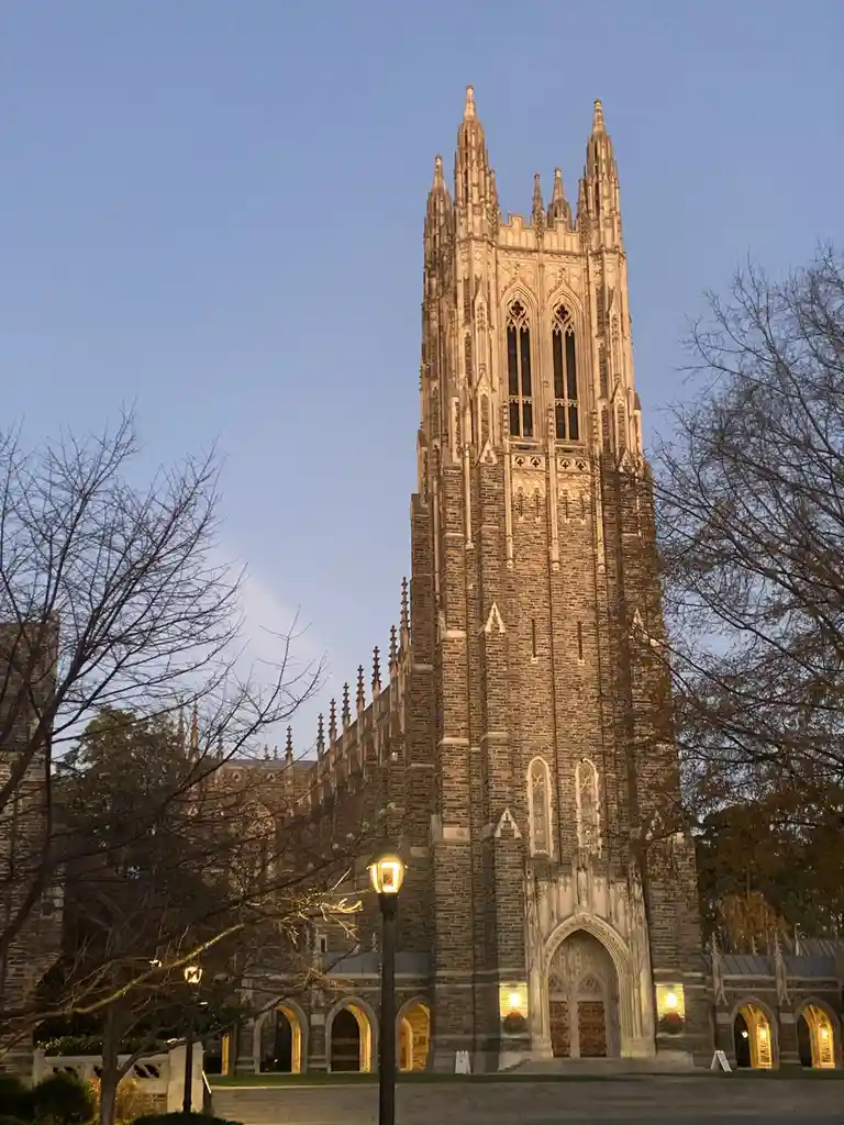A tall, ornate Gothic-style stone tower, likely a university chapel or cathedral, is illuminated at dusk against a clear…