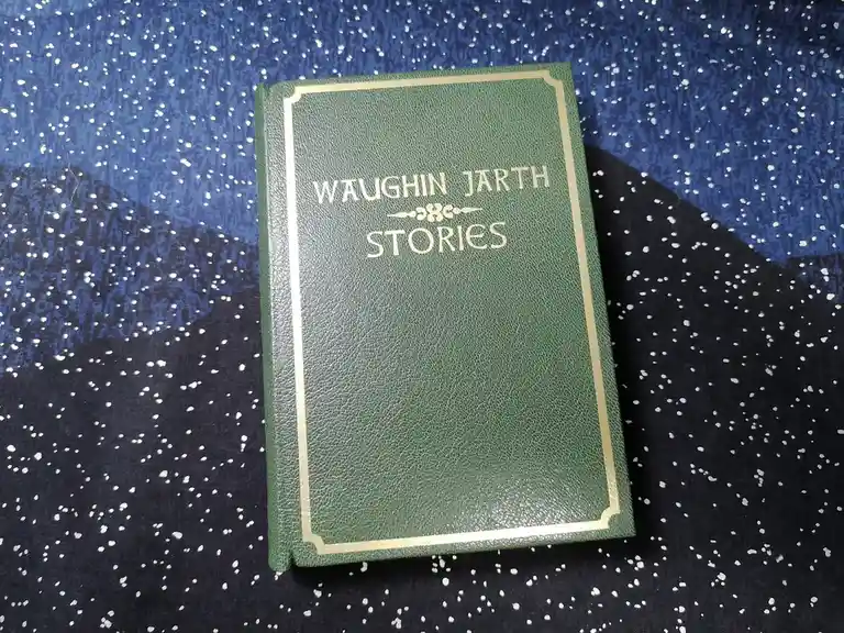 A green hardcover book with gold lettering reading 'WAUGHIN JARTH STORIES' sits on a blue and black fabric with white…