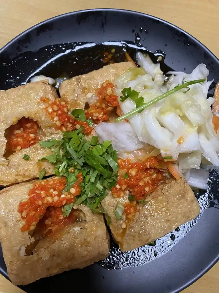A close-up photo of a black bowl containing fried tofu pieces topped with red chili paste, chopped cilantro, and shredded…