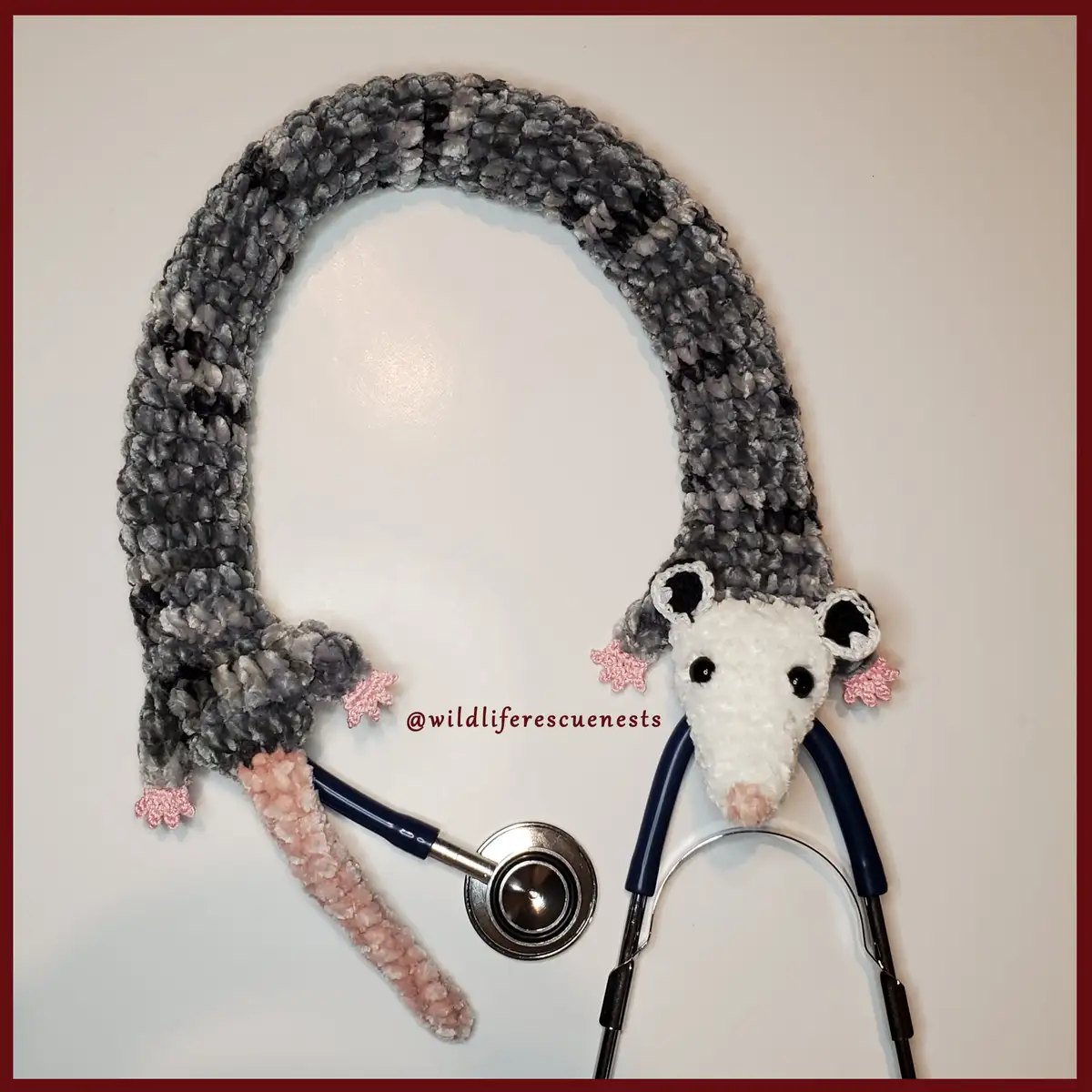 A crocheted opossum-shaped stethoscope cover with gray body, white face, black eyes, and pink paw details, draped over a…