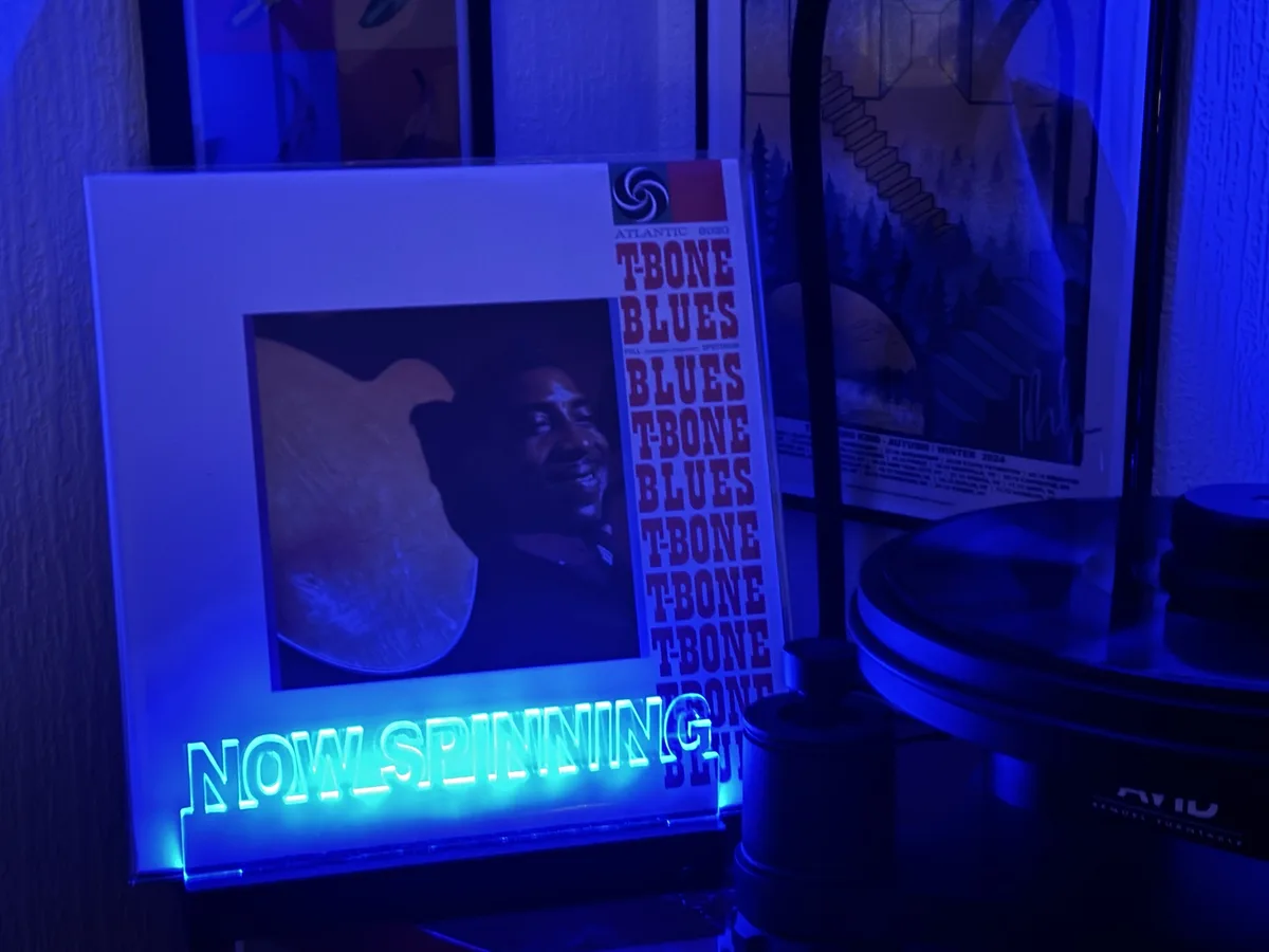 A vinyl record album titled 'T-Bone Blues' by Atlantic Records is displayed upright next to a turntable under blue lighting…