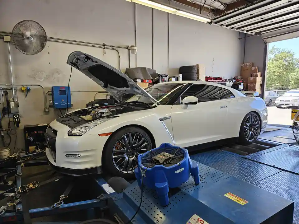 A white Nissan GT-R is parked on a blue car lift in a garage with its hood open, revealing the engine. In front of the car…