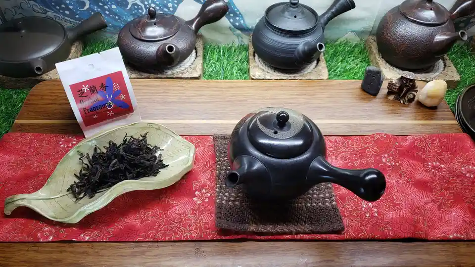 A still life of a traditional Chinese tea setup featuring a black clay teapot on a woven mat, next to a leaf-shaped dish…