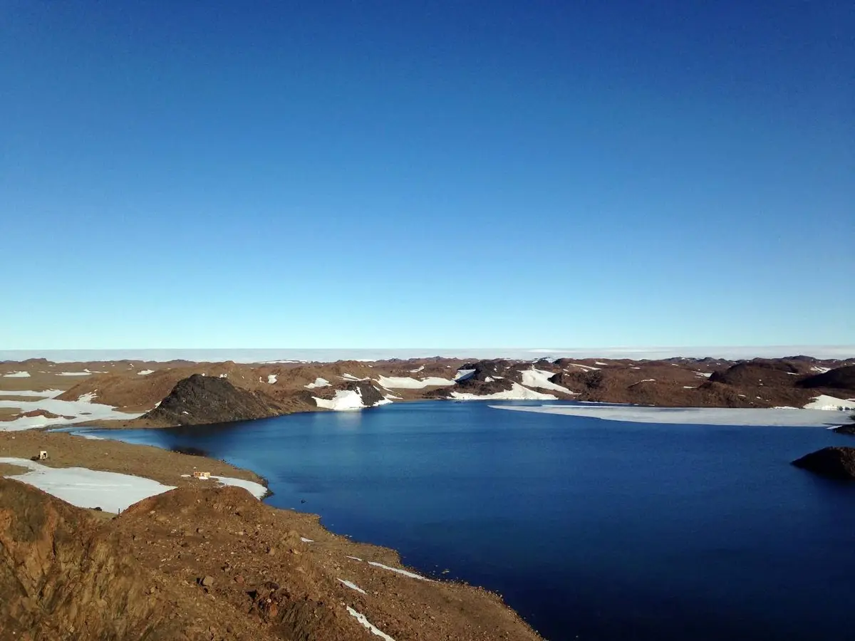 A wide-angle photo shows a vast, deep blue body of water surrounded by brown, rocky terrain with patches of snow, under a…