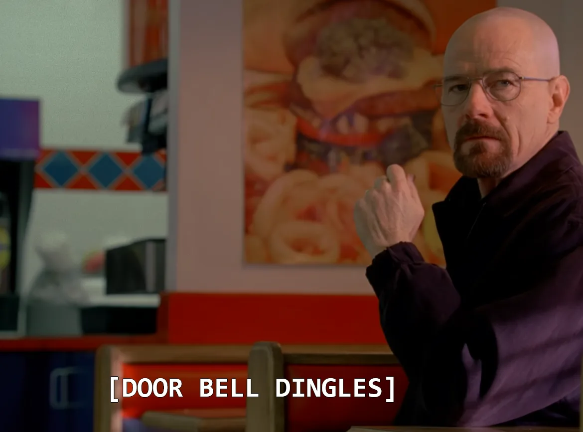 A bald man with glasses and a goatee, wearing a dark purple jacket, sits in a diner booth and looks intently forward with a…