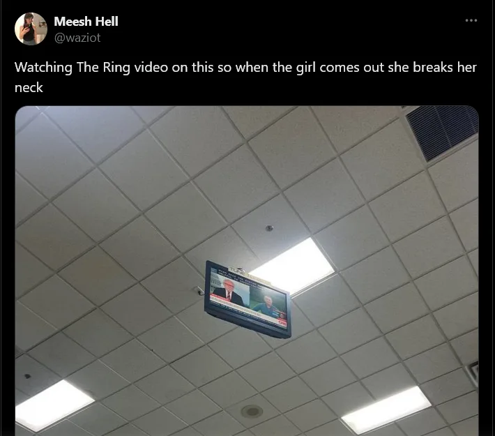 A photo of a small TV monitor mounted on a tiled ceiling in a public space, showing a news broadcast with two men. The…