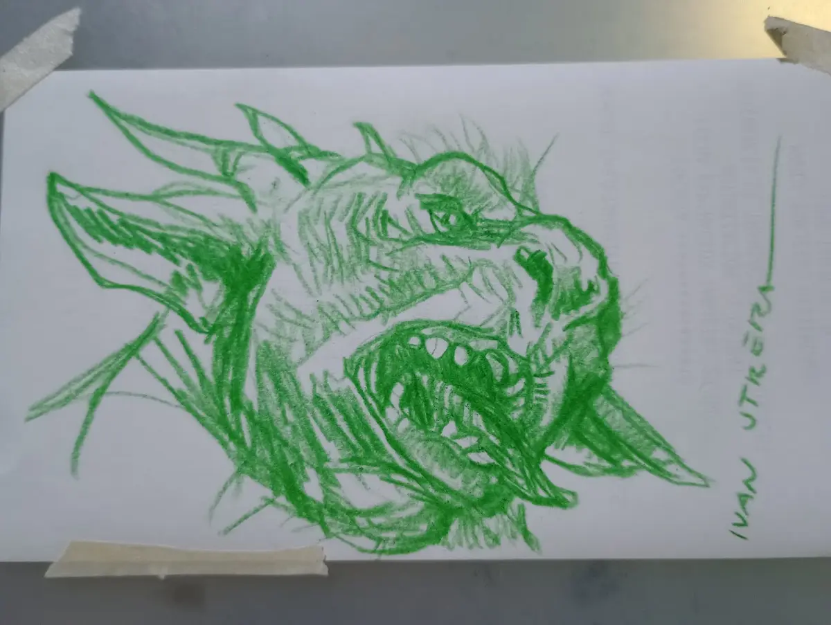 A hand-drawn sketch in green crayon on white paper depicts a fierce, snarling dragon-like creature with sharp teeth, spiky…