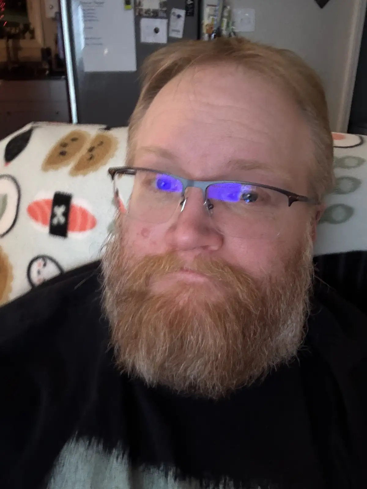 A man with a reddish beard and glasses, looking directly at the camera with a neutral expression, sits on a couch with a…