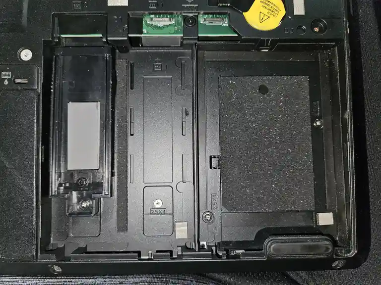 The image shows the inside of a laptop's bottom casing, revealing empty slots for a hard drive and optical drive, with a…