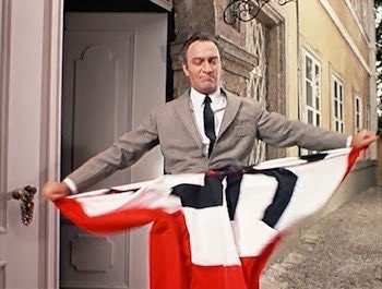 A man in a grey suit and tie stands in a doorway holding a large red, white, and black flag horizontally, looking directly…