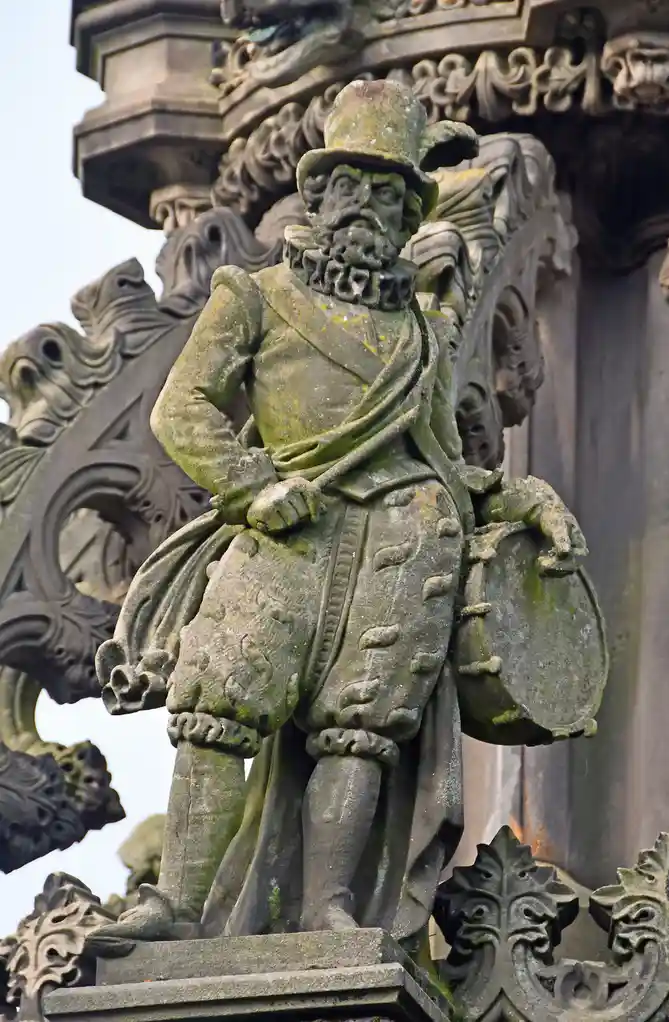 A weathered stone statue of a man in historical attire, including a hat, ruffled collar, and breeches, holding a drum. The…