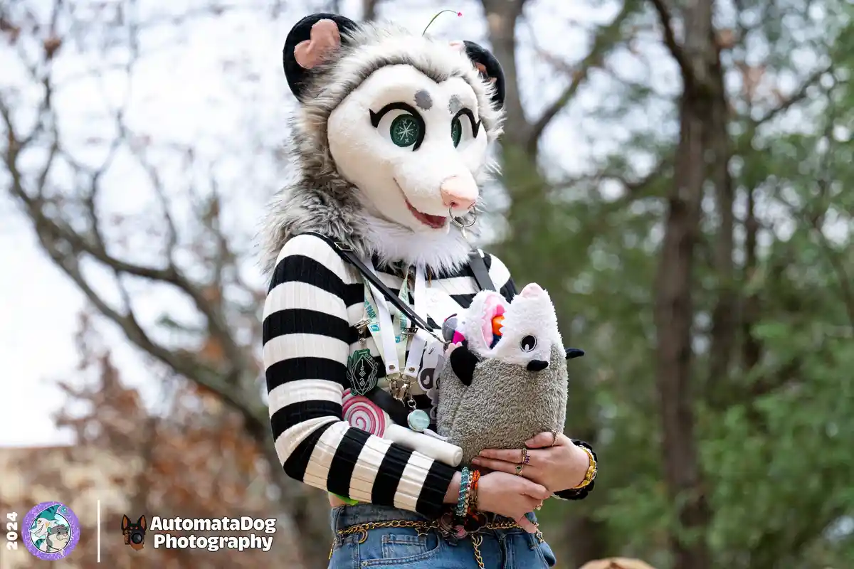 A person in a detailed, fluffy fursuit of an opossum with green eyes and a nose ring stands outdoors holding a plush toy…