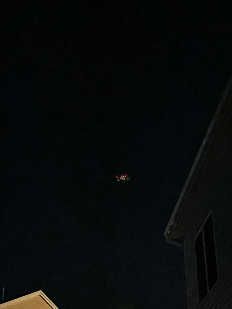 A small drone with red and green lights is flying in the dark night sky above rooftops of houses. The image captures the…