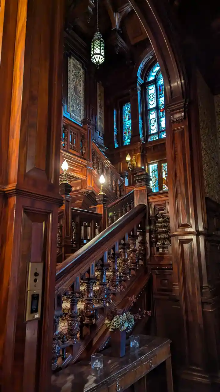 A grand, ornate wooden staircase with stained glass windows and intricate carvings is shown, with a modern elevator button…