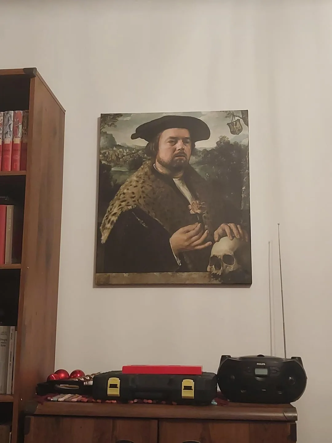 A painting of a man in historical attire holding a skull hangs on a wall above a wooden cabinet. The cabinet holds a black…