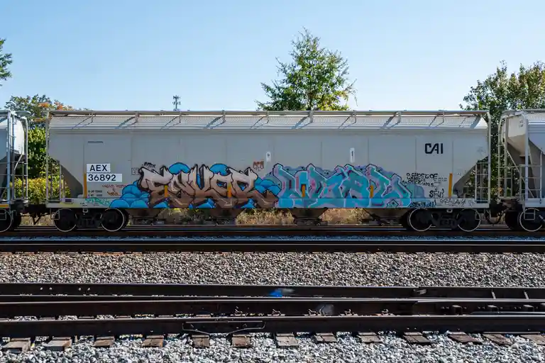 A side view of a white freight train car covered in colorful graffiti, sitting on railway tracks under a clear blue sky with…