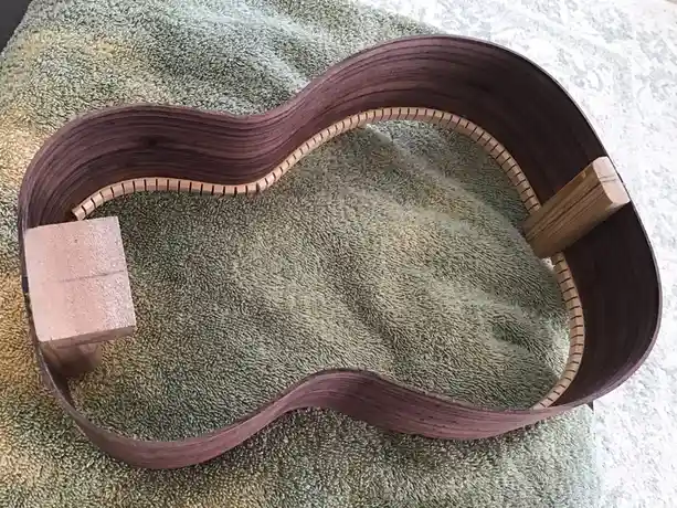 A handmade guitar body made of dark wood with a wavy, hollowed-out shape rests on a green towel. The inside edge has a…