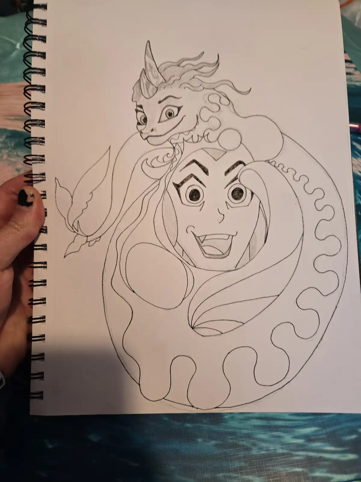 A hand holds a spiral-bound sketchbook displaying a pencil drawing of a cartoonish, smiling mermaid with a large, coiled…