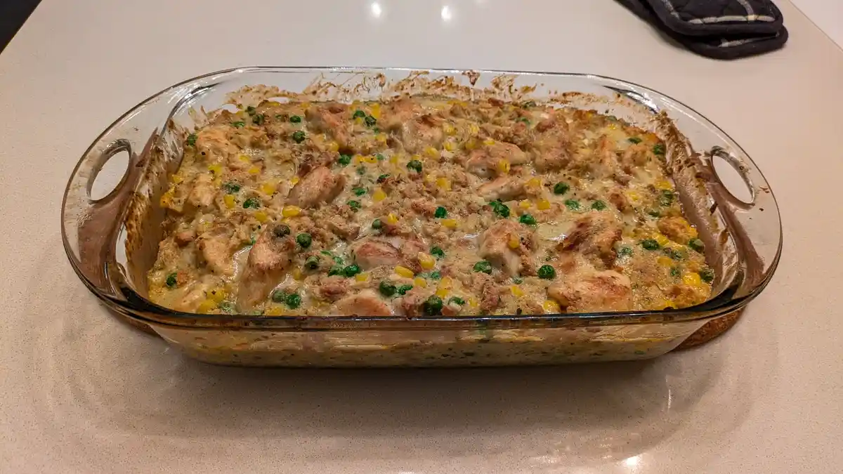 A freshly baked casserole in a clear glass baking dish sits on a light-colored countertop, featuring chunks of chicken…