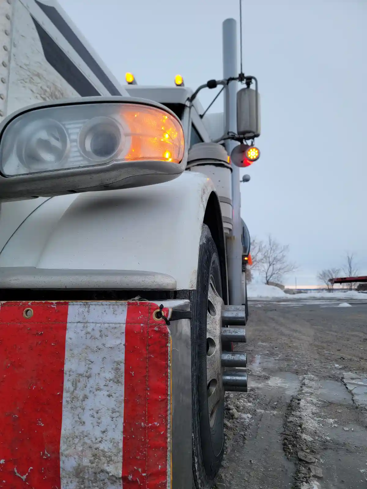 A close-up of the front-left side of a white semi-truck parked on a snowy, muddy roadside. The truck's headlight is on, and…