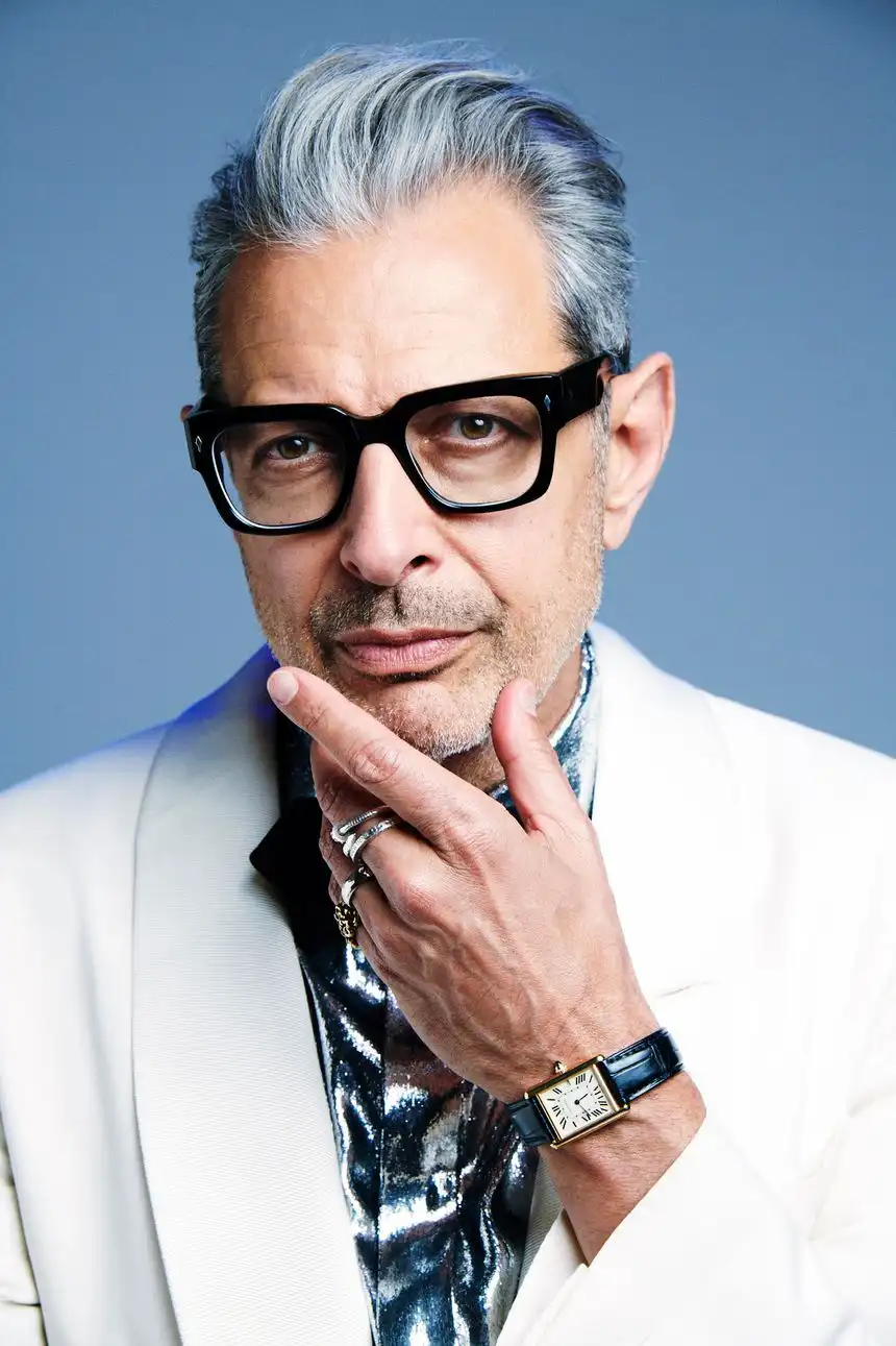 a man with gray hair and black-rimmed glasses wears a white suit jacket over a shiny silver shirt, his hand resting near his…