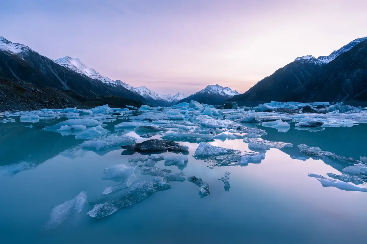 A serene landscape photo shows a calm, blue glacial lake filled with floating ice chunks, surrounded by snow-capped…