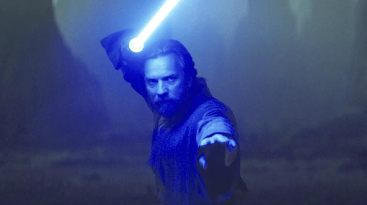 A man with a beard and hooded robe holds a glowing blue lightsaber above his head in a misty, dimly lit environment. The…